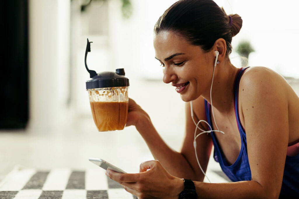Marcas Reprovadas de whey, Mulher de fone de ouvido sorrindo segurando o celular, na outra mão seguando a coqueteleira de whey